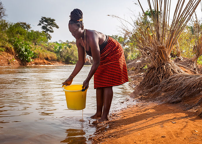 Woman collects water in yellow plastic bucket at a river.